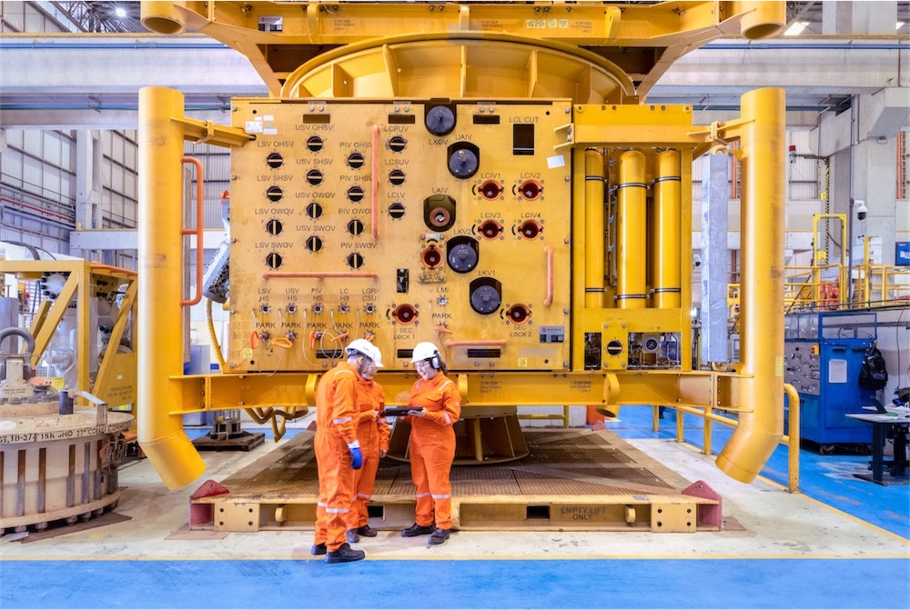 Three OneSubsea workers wearing orange jumpsuits standing in front of a subsea christmas tree discussing something.