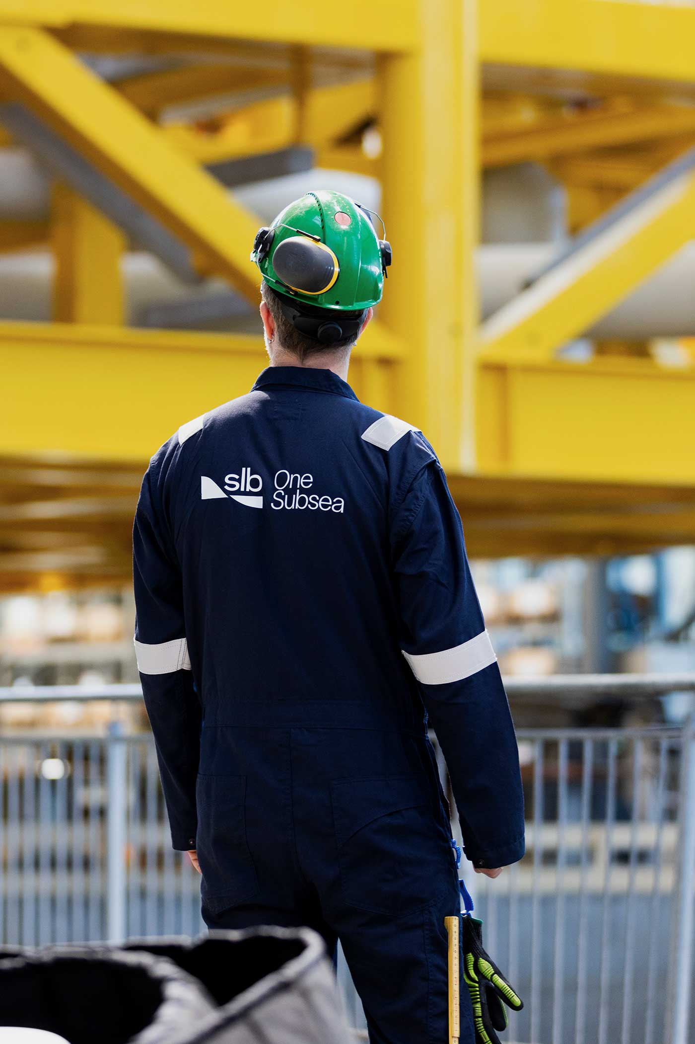 Industrial worker wearing a green hard hat and navy SLB OneSubsea coveralls walking through a large facility with yellow steel structures