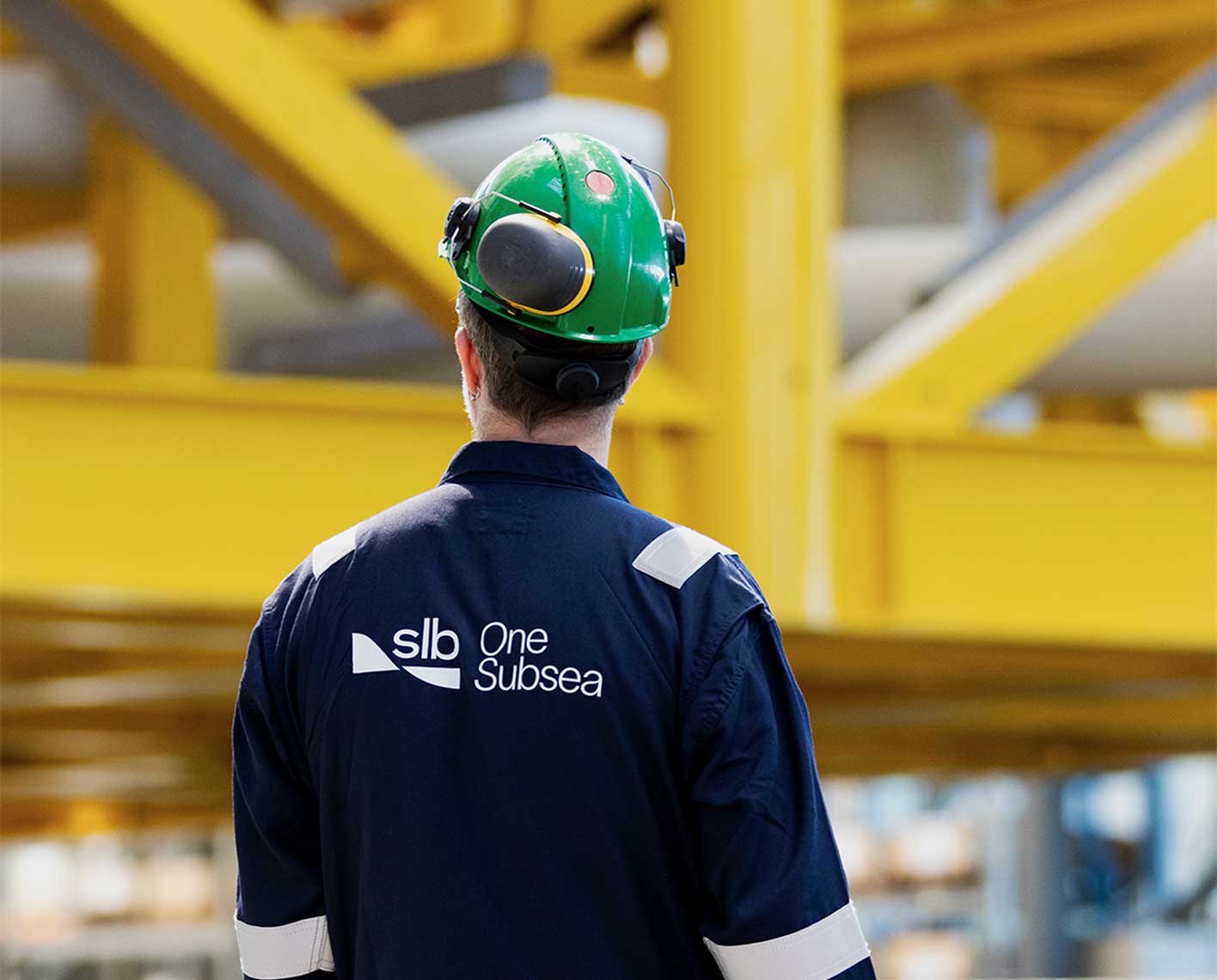 Industrial worker wearing a green hard hat and navy SLB OneSubsea coveralls walking through a large facility with yellow steel structures