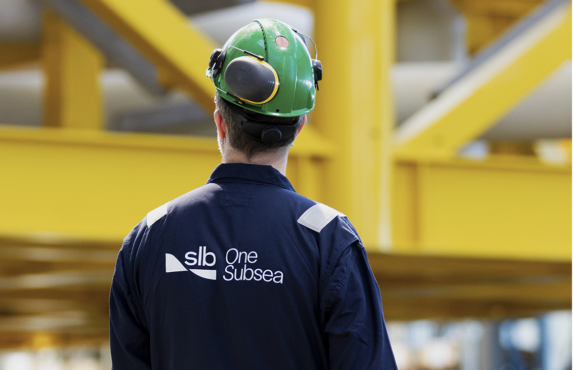 Industrial worker wearing a green hard hat and navy SLB OneSubsea coveralls walking through a large facility with yellow steel structures