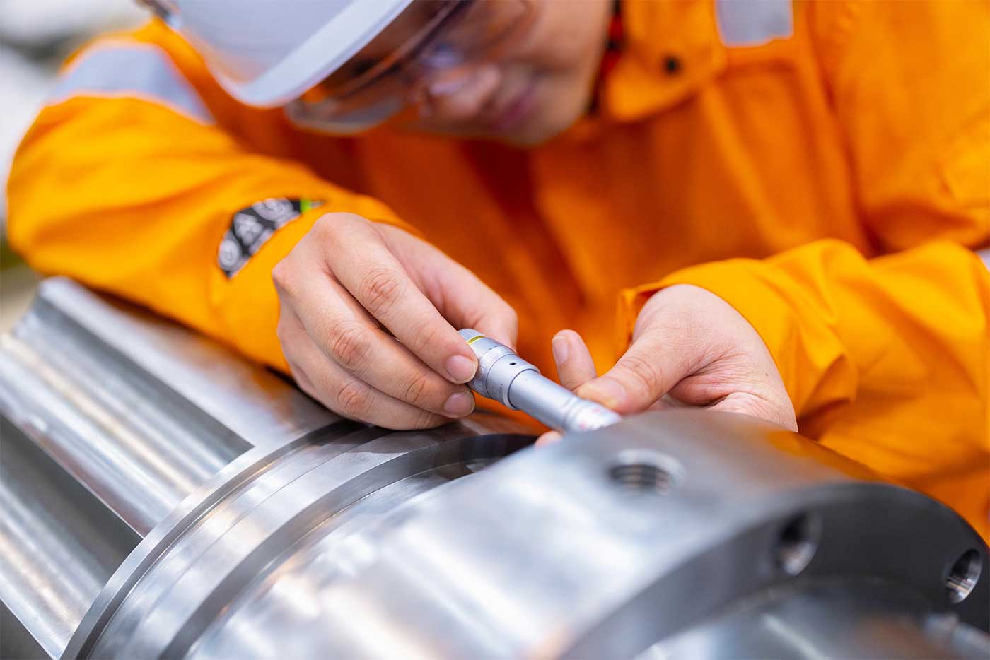 Close-up of a worker in orange safety gear and helmet using a precision measuring tool to inspect a metal component in an industrial setting.